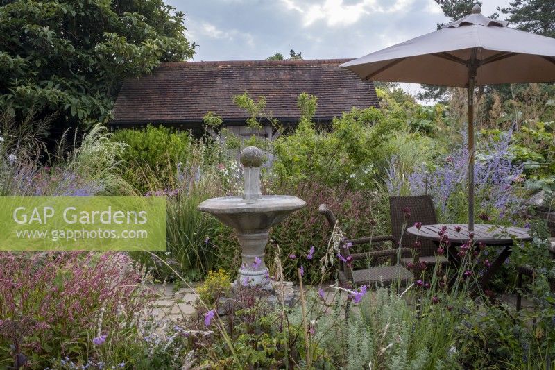 Small courtyard garden filled with late summer planting, with framed oak building behind and water feature, bird bath and table with umbrella in foreground