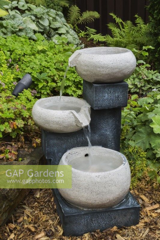 Cascading granite bowls water fountain in mulch border in backyard garden in summer.