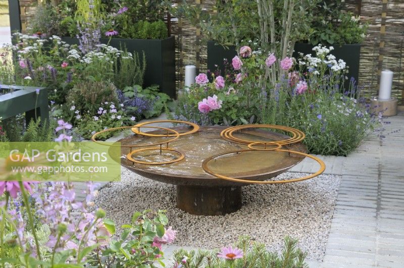 A weathered mild steel water feature with rings set in a gravel square surrounded by a mixture of planting influenced by aromatic and wildlife-friendly ornamentals in a design structure inspired by the parterre.  Rosa 'Princess Alexandra of Kent' in the background.  Landform Mental Wealth Garden - RHS Hampton Court Palace Garden Festival 2023. 