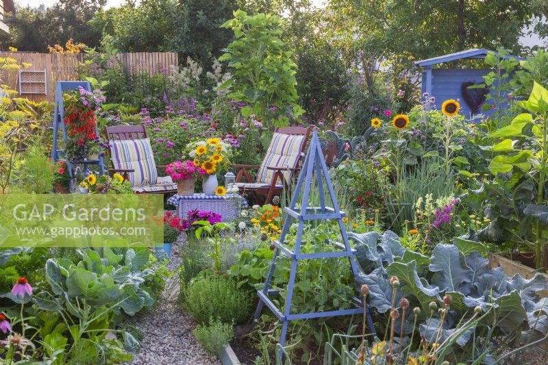 In front are vegetable beds with growing vegetables, and in the back garden furniture with a flower arrangement, a wooden gazebo and a bed with perennials.