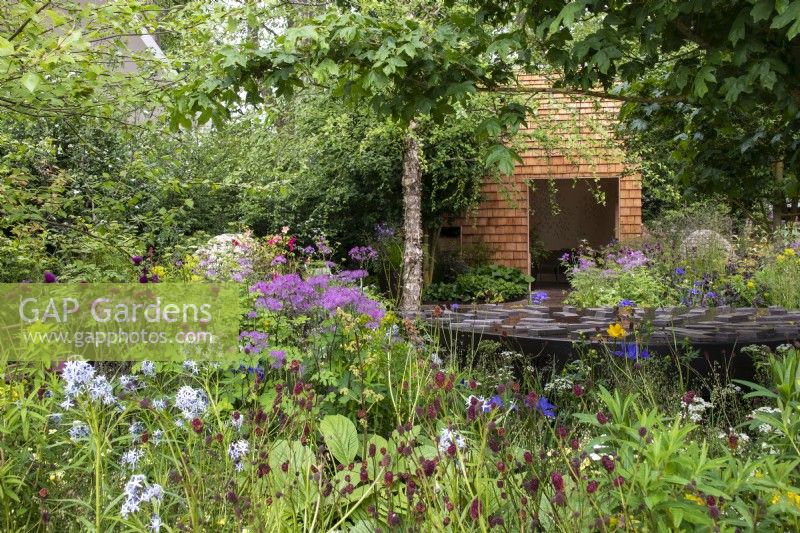 View across borders and an interactive water feature to the woodland retreat on the wheelchair accessible Horatio's Garden - Designers Hugo Bugg and Charlotte Harris, Harris Bugg Studio - RHS Chelsea Flower Show 2023