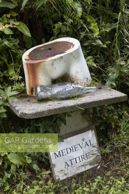 An old mannequin torso is on a plinth with a glass fish shaped bottle. A sign saying 'Medieval attire' is below. The sculpture puzzle represents cod piece. Harbour Lights, Devon NGS garden. July. 