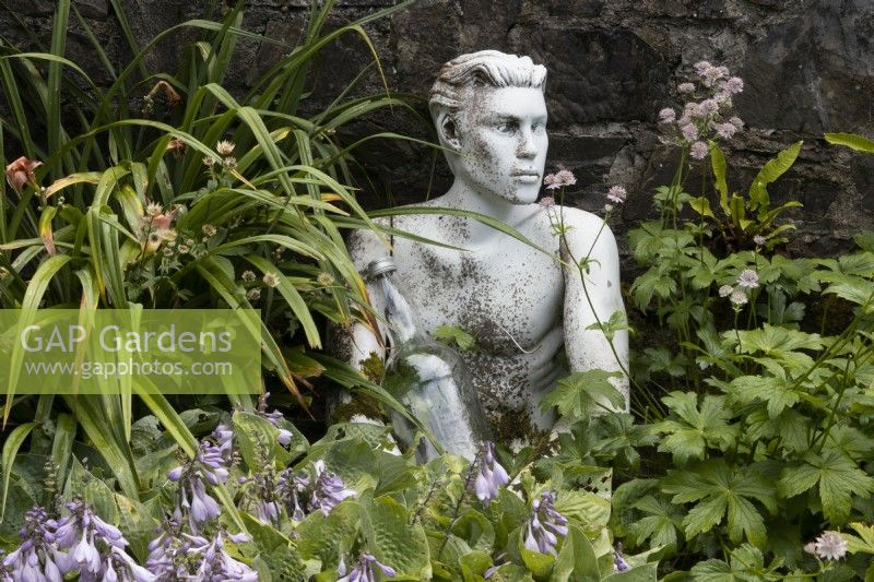 An old mannequin head and torso sit at the base of a wall amongst a variety of foliage and flowers. An old bottle is held by the mannequin. The sculpture puzzle represents 'Legless'. Harbour Lights, Devon NGS garden. July. 