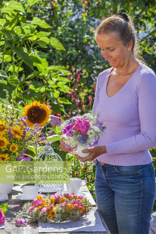 Woman arranging outdoor table with wreath and bouquets of flowers in vases. Flowers in bouquets are Hydrangea, sweet peas, Ammi majus and Nigella while wreath is made of Clematis seed heads, Dahlia and Rudbeckia.