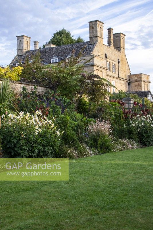 Early morning sun on flower border, lawn and house at Bourton House Garden, Gloucestershire.