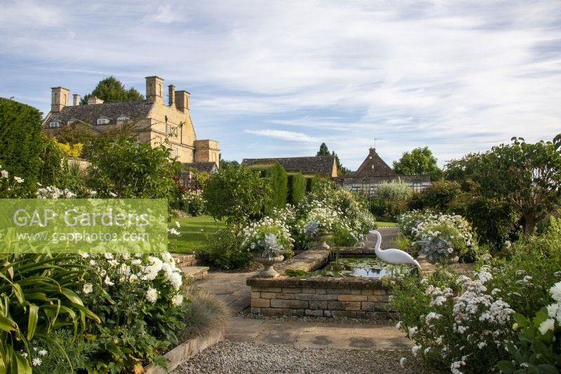 View across the White Garden with raised formal pond, and borders planted with perennials towards the house at Bourton House Garden, Gloucestershire.