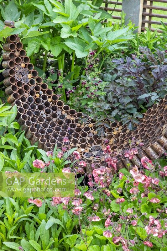 A rusty garden sculpture made from reclaimed, recycled materials with spaces made for an insect hotel - mixed planting of Geum 'Pink Petticoats', Geranium phaeum 'Samobor', Angelica sylvestris 'Vicars Mead' - syn. 'Vicar's Mead'