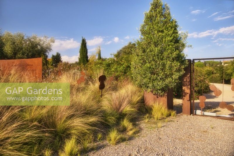 Mediterranean  garden view at the entrance gates decorated with musician instruments elements, grassland with Stipa tenuissima, Olive tree in the iron container and violin sculptures in autumn time, sunlit with warm evening light.

Italy, Tuscan Maremma, Orbetello
Autumn season, October
