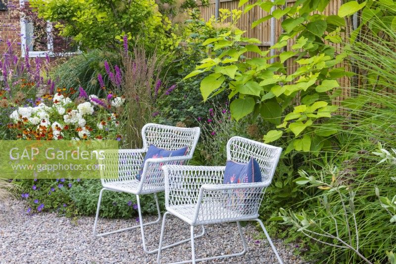 In the middle of the garden, two wirework chairs sit on a pebbled area, set against a backdrop of helenium, lythrum, white hydrangea and hardy geranium.