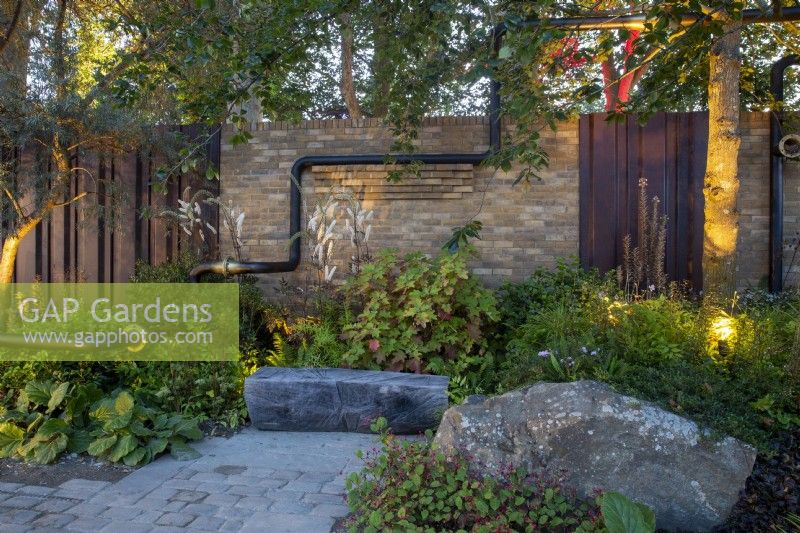 Urban garden lit up at night reclaimed cobble stone paving - mixed perennial planting of Hydrangea quercifolia, Actaea simplex 'Atropurpurea' and Persicaria 'Indian Summer' - carved tree trunk bench seating area - brick and metal panel wall