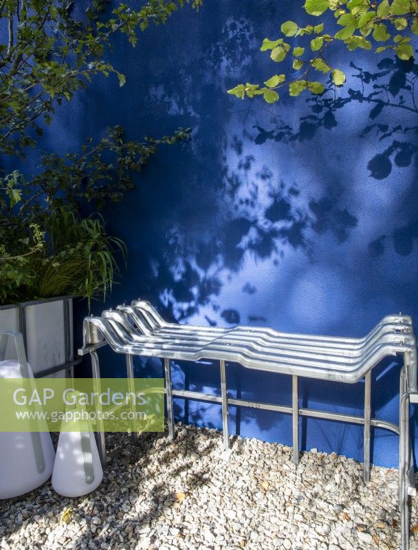 Repurposed upcycled modern contemporary metal bench seat made from an IBC - intermediate bulk container frame - on a gravel surface patio - blue painted rendered wall