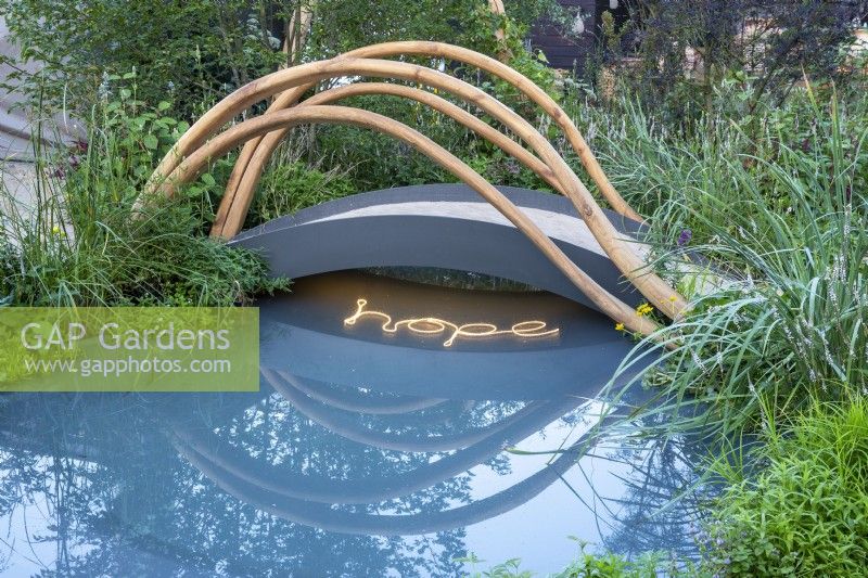 A modern contemporary small metal arched bridge with sculptural wood rails over a pond with HOPE written in lighting in the water - water marginals