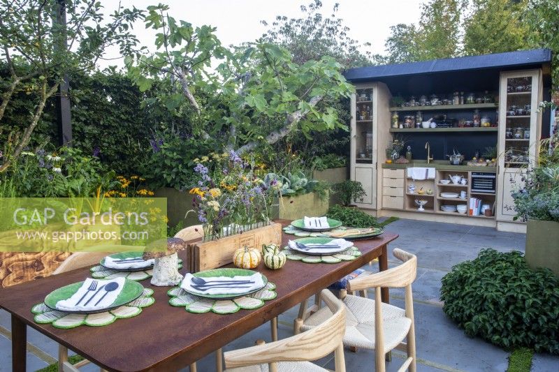 An outdoor kitchen with a paved patio area with dining table. A Ficus carica - fig tree and charred wood wooden raised beds with mixed perennial edible planting - 