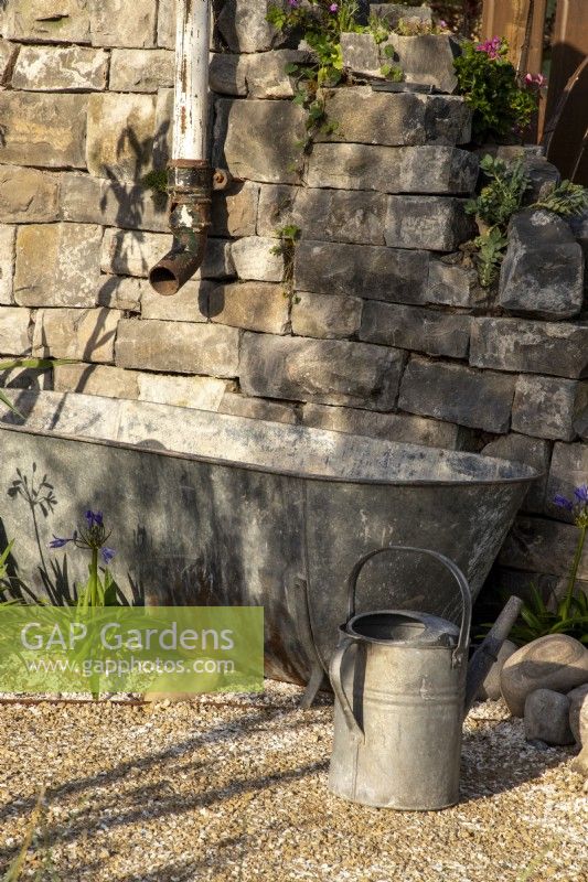 A drystone wall with a metal drainpipe - an old rustic galvanised metal bath tub water butt for collecting rainwater - galvanised metal watering can on a crushed shell and gravel surface path