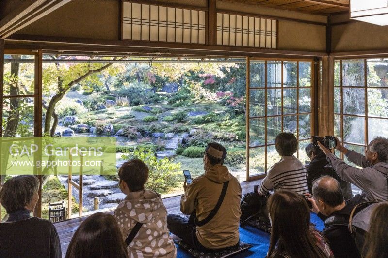 Group of visitors sitting in main building taking pictures and looking at view into garden framed by sliding glass walls. 
