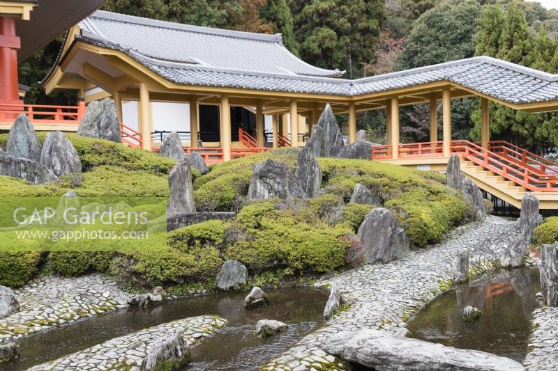 View across paved area with small pools and placed rocks with bed of Rhododendron (not in flower). Steps and pavillion of temple building. 