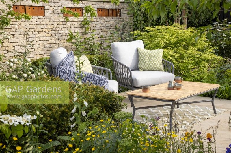 A patio seating area with table and chairs - Taxus baccata hedges, mixed perennial planting Geranium phaeum 'Raven', Ranunculus acris - drystone wall with corten steel bird boxes