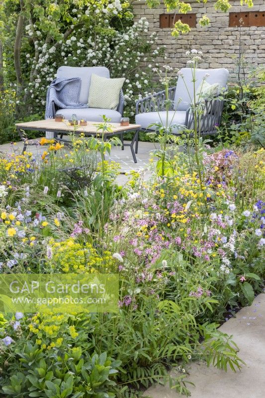 Patio seating area with table and chairs, mixed perennial planting border Geranium pratense 'Mrs Kendall Clark', Lychnis flos-cuculi, Euphorbia robbiae, Sanguisorba 'Alba', Ranunculus acris - drystone wall with corten steel nesting boxes 