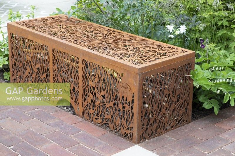 A laser cut corten steel dead hedge for filling with garden prunings that create habitats for beneficial insects and entrance for hedgehog boxes - next to a brick paved path