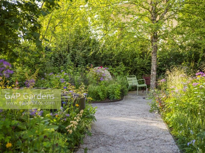 Path surrounded by flowering borders with water feature leading to seating area Fernob chairs, Betula nigra, stone cairns at Horatio's Garden, Chelsea Flower Show 2023. Designer: Charlotte Harris and Hugo Bugg, Gold medal winner, Best Show Garden