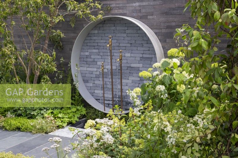 Garden wall with a circular concrete and clay brick pavers water feature, brass tap spouts and copper pipes, mixed perennial planting in white and green tones 