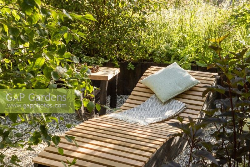 A wooden recliner with cushion and blanket and a small table in a woodland garden mixed perennial planting and ornamental grasses