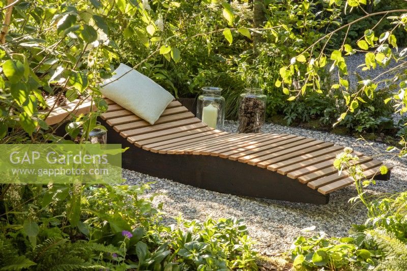 A wooden recliner with cushion in a woodland garden with mixed perennial planting 