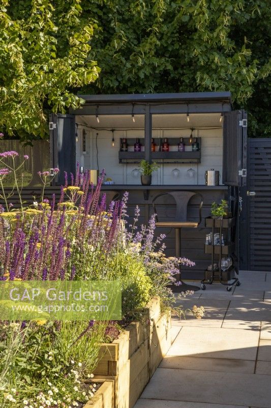 A raised timber bed on a patio with planting of Salvia nemorosa 'Caradonna', Achillea 'Terracotta' and Verbena bonariensis in front of an outdoor bar cabin