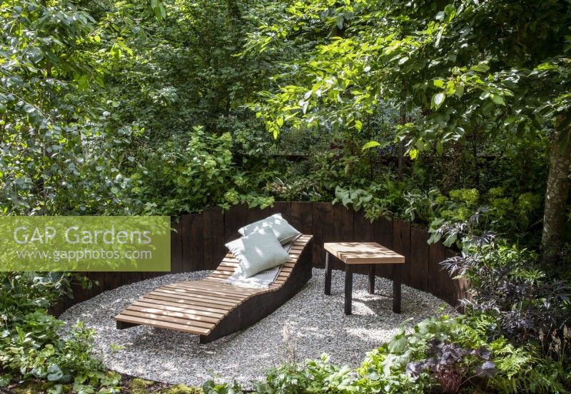 An enclosed relaxing area with gravel surface and wooden raised beds,  wooden recliner with blanket and cushions in a woodland garden with mixed perennial planting 