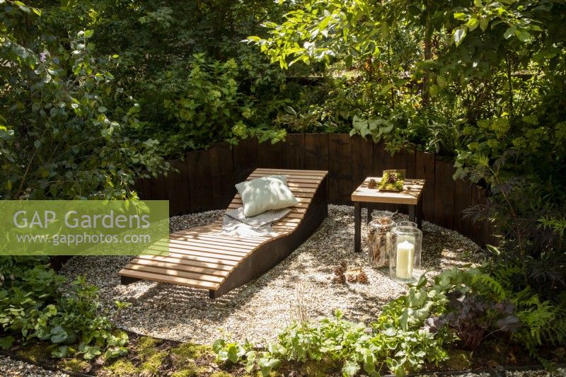 An enclosed relaxing area with gravel surface and wooden raised beds,  wooden recliner with blanket and cushion in a woodland garden with mixed perennial planting 