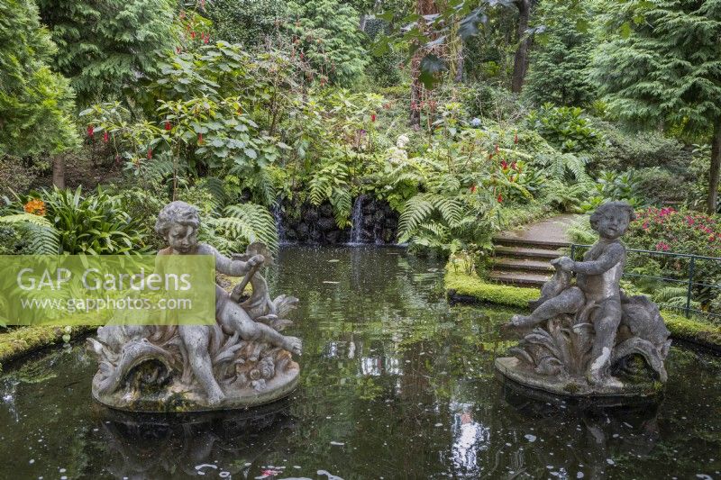 Two figurative statues in a pond, with small waterfalls in the background, steps to the right and lush tropical planting in the background. Monte Palace Gardens, Madeira