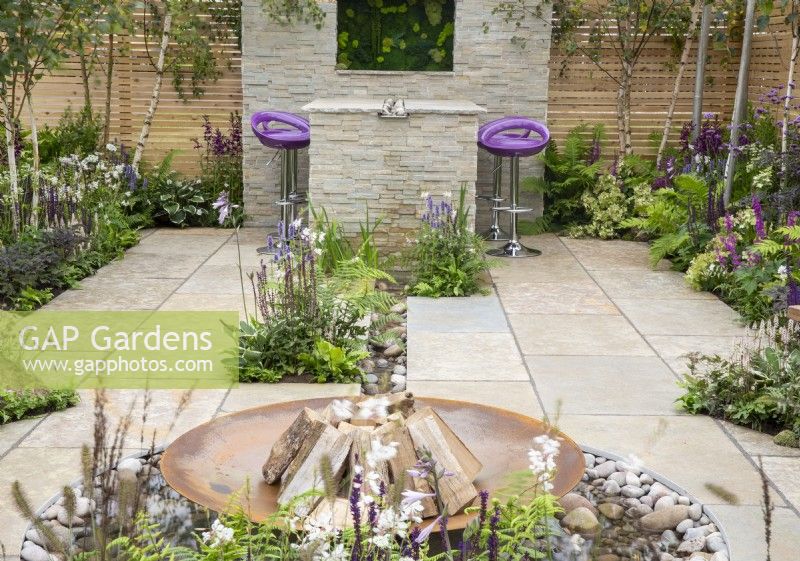 Stone paved patio with a corten steel fire bowl, dining area with purple bar stools, mixed perennial planting in purple and white colours