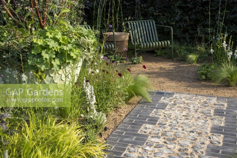 A cobbled stone flint and grey brick path leading to a gravel surface seating area and mixed perennial planting border including Cosmos atrosanguineus