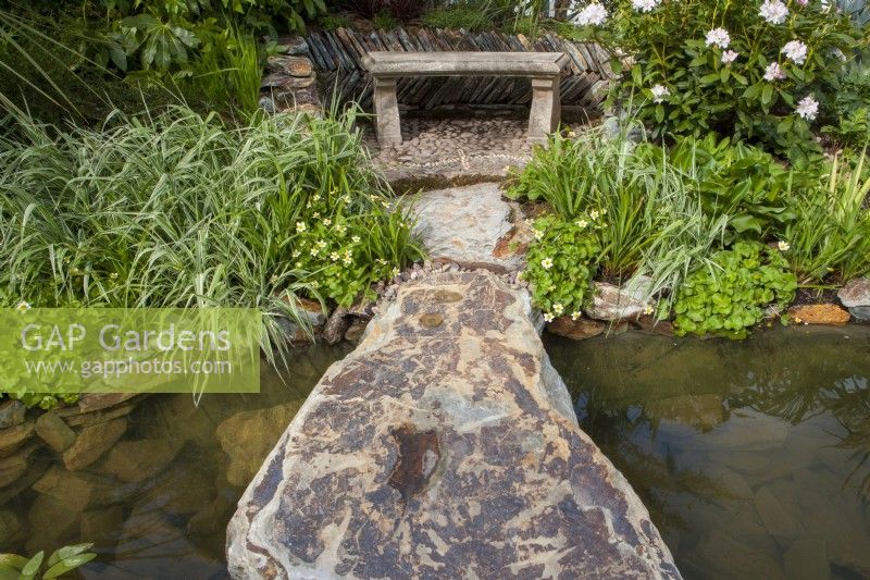 Boulder over a pond leading to a stone bench with drystone wall surround, marginal planting of Phalaris arundinacea and Rhododendron