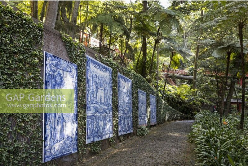 Antique tiled panels adorn an ivy covered wall with trees overhead and a cobbled path in front. Monte Palace Gardens, Madeira. August. Summer