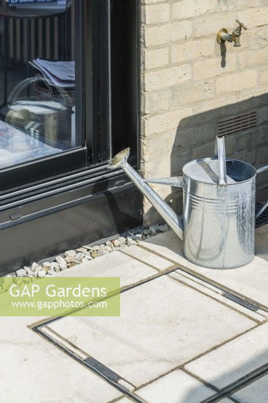 Close-up of drainage inspection cover with inset limestone paving next to patio doors. Outside tap with galvanised watering can. June