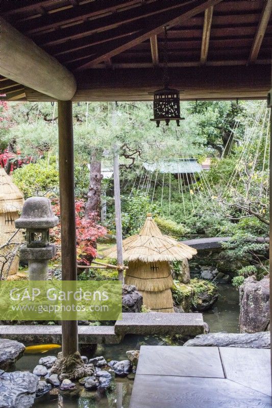 View from veranda to stone bridge crossing water with straw-wrapped fragile stone ornament as protection against frost. Shrubs with pole and rope protection against snow damage called Yukitsuri. Stone lantern called Ishidoro.
