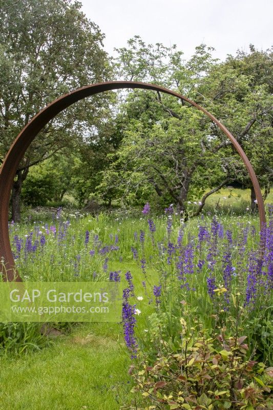 A mown path through a Corten steel moon gate in a perennial wildflower meadow.