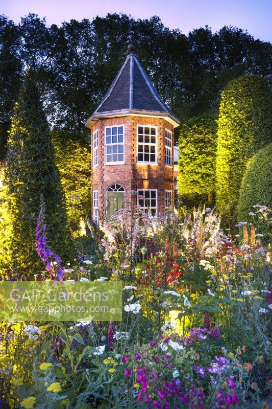 Highly colourful herbaceous border and evergreen topiary leads to a small brick house lit at night. Plants include Eremurus 'Pinocchio', Digitalis purpurera - foxglove, lupins, Verbena bonariensis, pink roses, and a bay topiary cone.

The Harrods British Eccentrics Garden for RHS Chelsea Flower Show 2016 designed by Diarmuid Gavin.