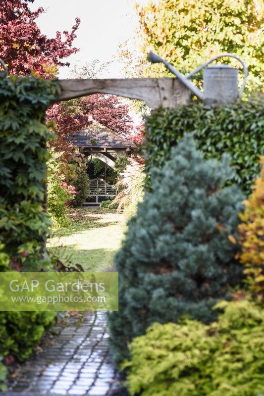 Path leading beneath a carved oak entranceway leading into a lawned area of the garden towards a gazebo with seat in November