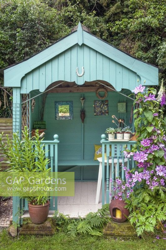 Turquoise painted garden summerhouse, wall ornaments, containers with Sempervivums and lilies, trained Clematis 'Nelly Moser' climber,  Digitalis purpurea - foxgloves, 
Geranium palmatum - Canary Island geranium and ceramic lamp for candles.