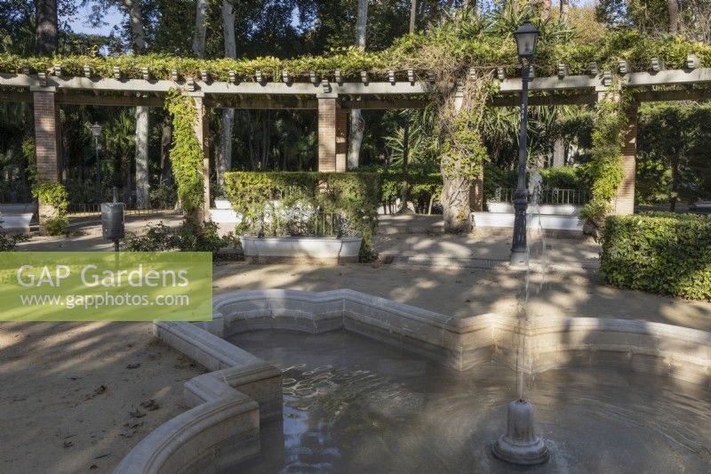 A stone fountain in front of a large curving pergola. Parque de Maria Luisa, Seville, Spain. September