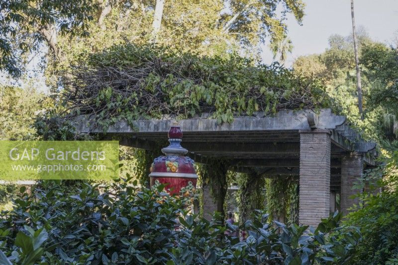 A colourful glazed urn stands in between a hedge and a large pergola in the Parque de Maria Luisa, Seville, Spain. September