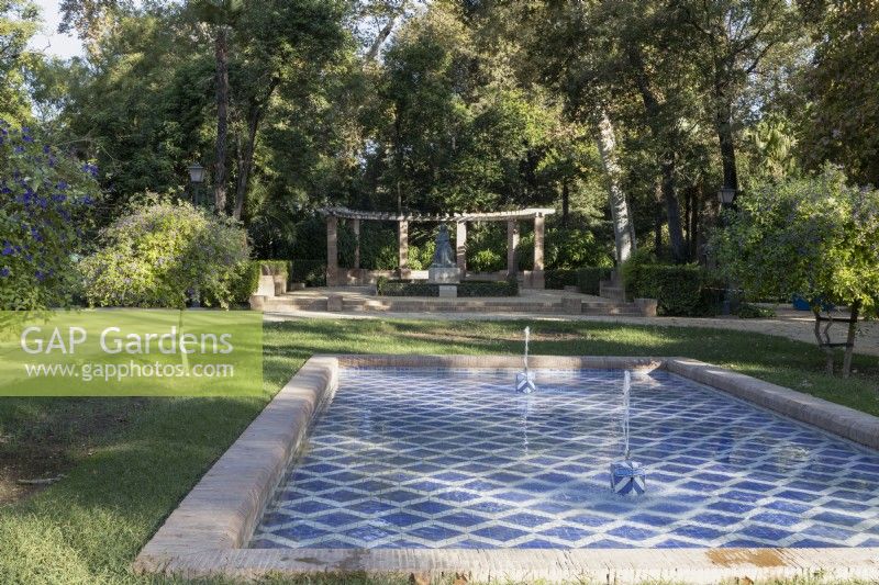 A blue and white tiled pool with two fountains' large curved pergola is in the background. Parque de Maria Luisa, Seville, Spain. September