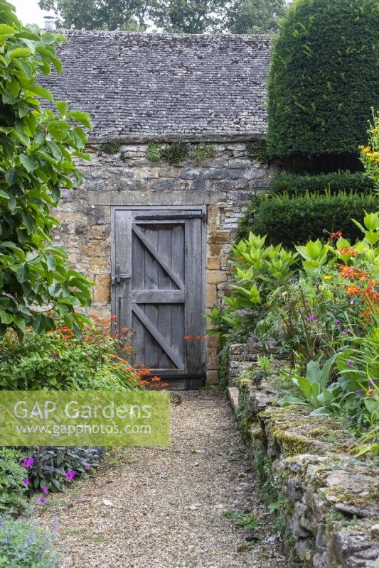 A gravel path leads past a Cotswold stone retaining wall, yew topiary and summer flowers towards a wooden door in a brick wall at The Manor, Little Compton.