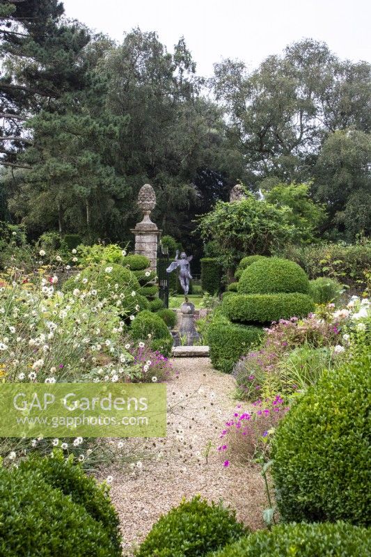 A view through The Flower Garden towards the pond and fountain with a gravel path, perennials and box topiary.