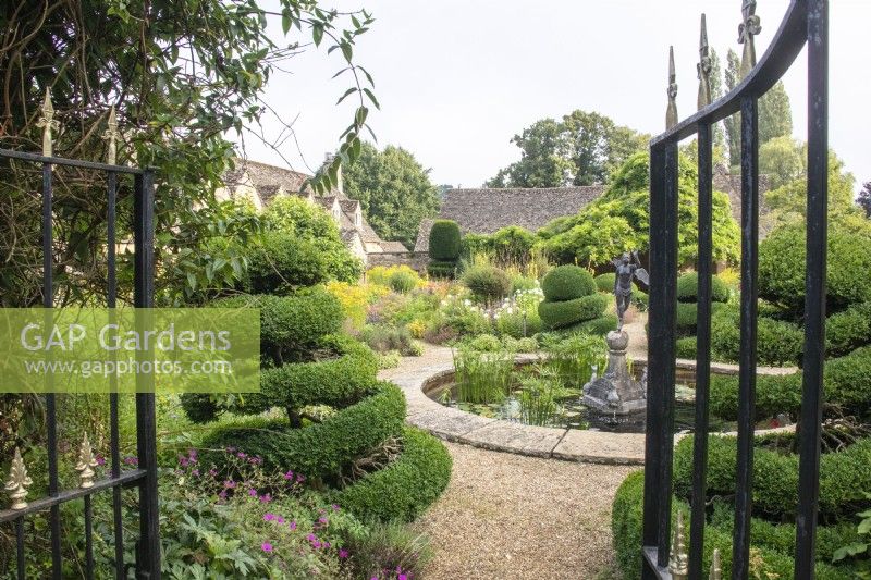 A view through gates into The Flower Garden with perennials, topiary and a circular pond at The Manor, Little Compton.