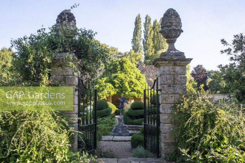 Wrought iron gates and stone pillars with decorative finials frame a view across The Flower Garden towards a summer house at The Manor, Little Compton.