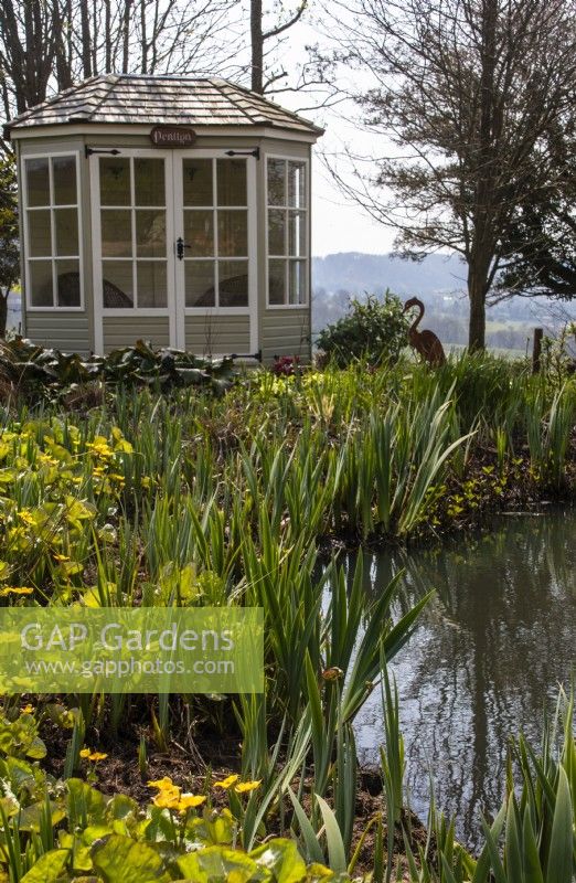 A hexagonal wooden summerhouse alongside a wildlife pond planted with marginals at Trench Hill, Gloucestershire