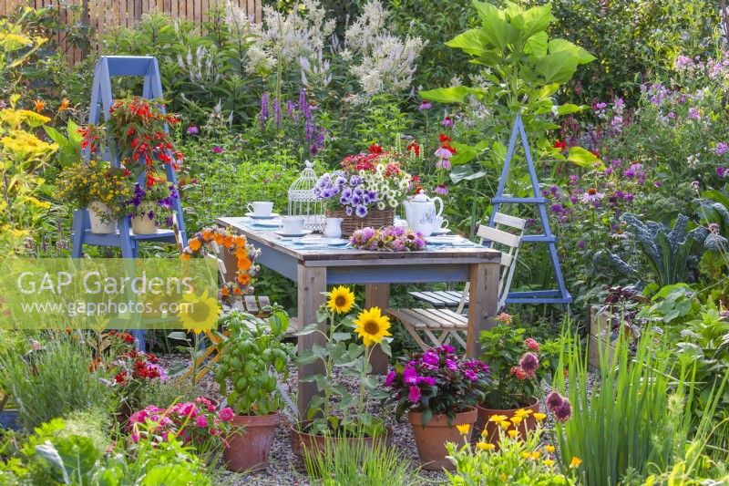 Outdoor dining area with table set for tea and containers planted with bedding flowers and herbs including Surfinia, Impatiens, basil, sunflowers, Verbena and others.